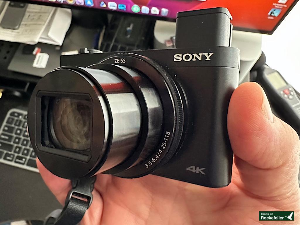 A person holds a black Sony 4K camera with an extended lens. A computer monitor, keyboard, and papers are visible in the background.