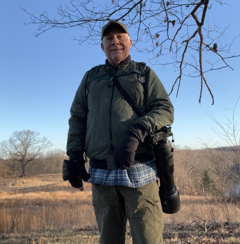 A person stands outdoors in a field holding a camera with a long lens. They are wearing a green jacket, cap, and gloves. Bare trees and a clear sky are in the background.