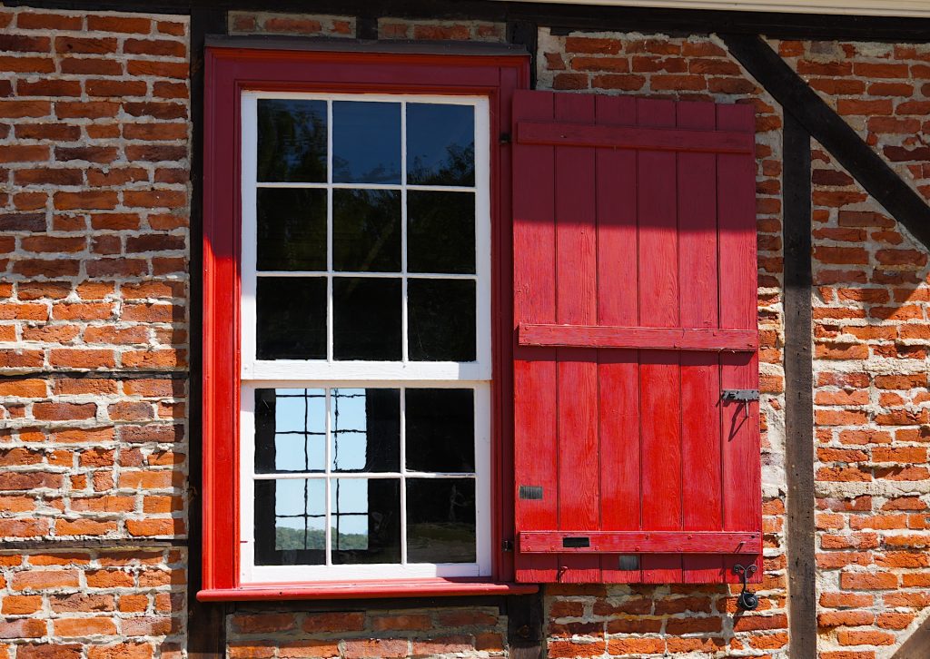 A red-framed window with a closed red shutter on a brick wall. The window has two sections with grid patterns, reflecting blue sky and dark tree silhouettes.