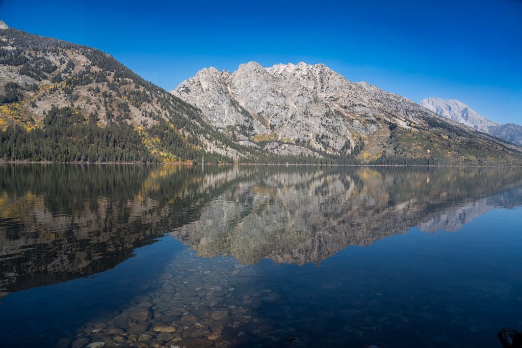 Mountain range reflected on a calm, clear lake surrounded by trees under a blue sky.