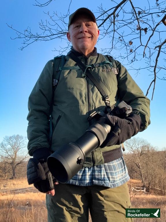 Man outdoors wearing a green jacket and gloves, holding a large camera lens. Brown landscape and bare trees in the background. "Birds of Rockefeller" logo in corner.