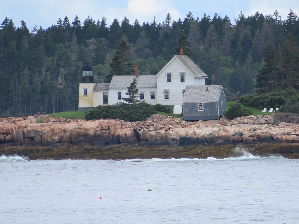 White house and lighthouse on a rocky coastline, surrounded by trees, with the ocean in the foreground.