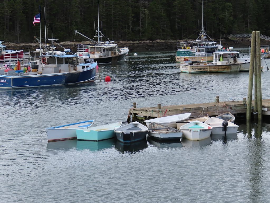 Small boats are moored alongside a wooden dock, with larger fishing boats floating in the background on a calm body of water.