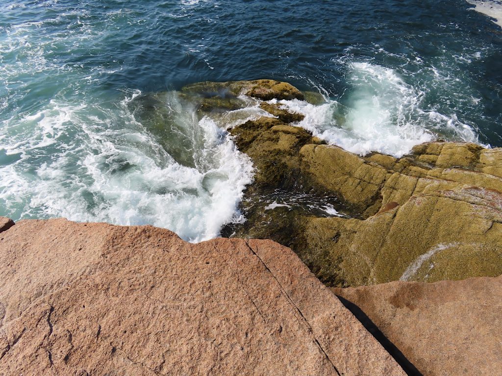 Waves crashing over rocky shoreline with clear blue water and light brown rocks.