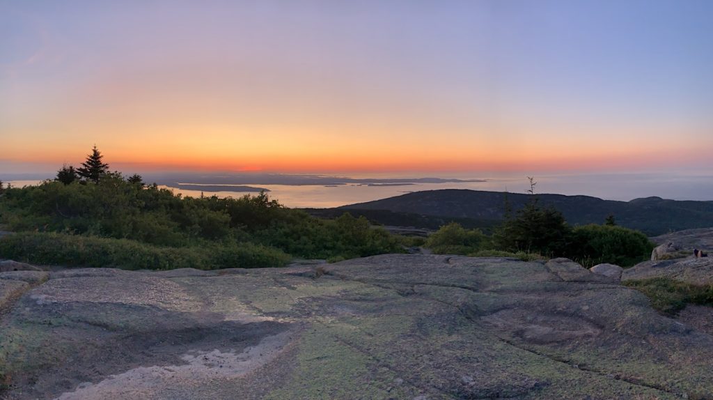 Sunrise over a rocky landscape with trees, overlooking a distant water body and islands under a clear sky.