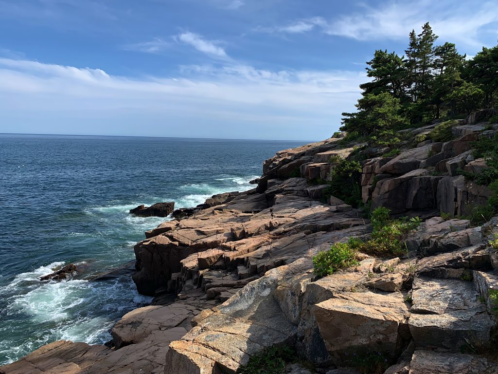 Rocky coastline with waves crashing against the shore under a blue sky, with green trees on the cliffs.