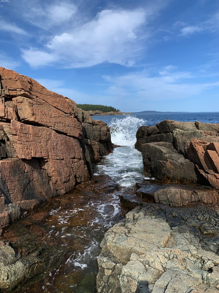 Rocky coastline with red and gray cliffs, a narrow inlet with water splashing through, and a distant view of the ocean under a partly cloudy blue sky.