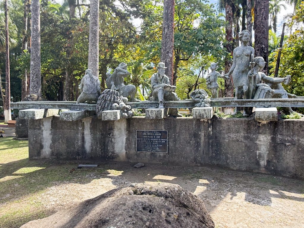 Bronze statue of workers and animals on a wagon, set on a concrete platform, surrounded by trees. Plaque is visible below the sculpture.