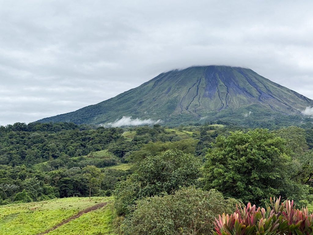 A view of a green, tree-covered landscape with a large, cloud-capped volcano in the background under a cloudy sky.