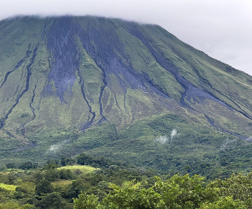 A lush green landscape with a towering, cloud-covered volcanic mountain in the background, featuring visible dark lava flows on its slopes.