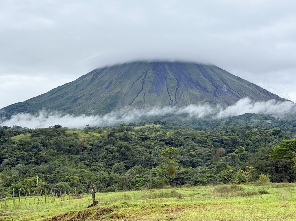 Cloud-covered volcano with green forested slopes under a cloudy sky.