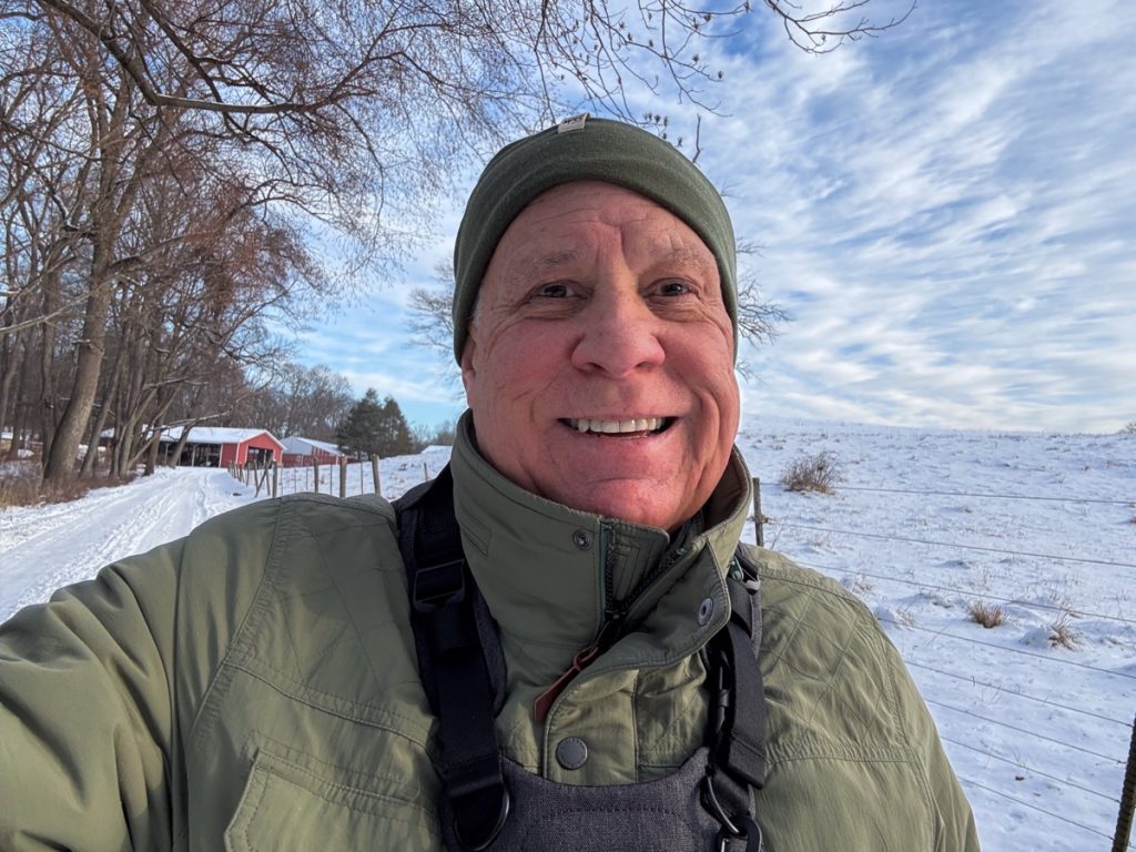 A person wearing a green jacket and beanie smiles while standing outside on a snow-covered path with a red barn and trees in the background.