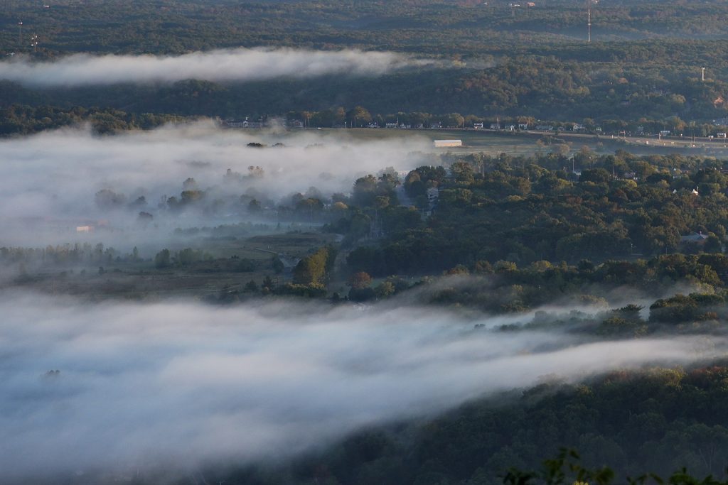 Aerial view of a lush green landscape partially covered with low, dense fog, revealing winding roads and scattered buildings beneath.