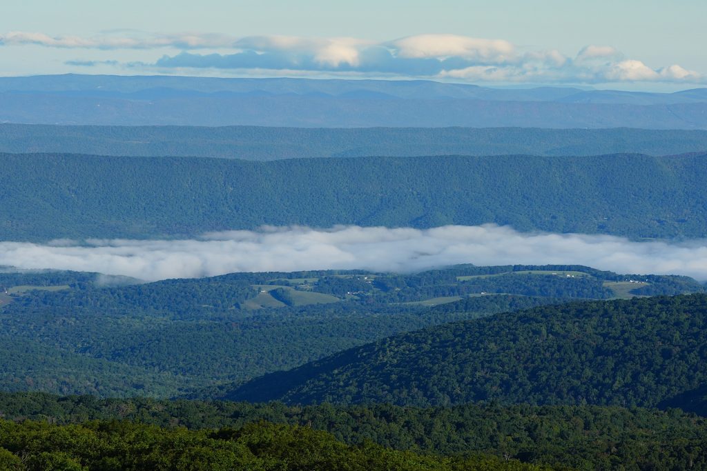 A scenic view of layered blue mountain ranges with fog settled in the valley and scattered clouds in the sky.