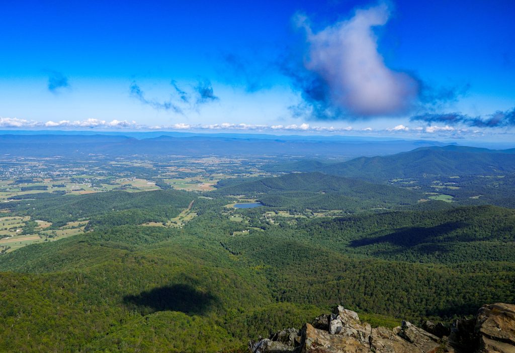 Aerial view of a vast landscape with forests, fields, and distant mountains under a blue sky with sparse clouds.