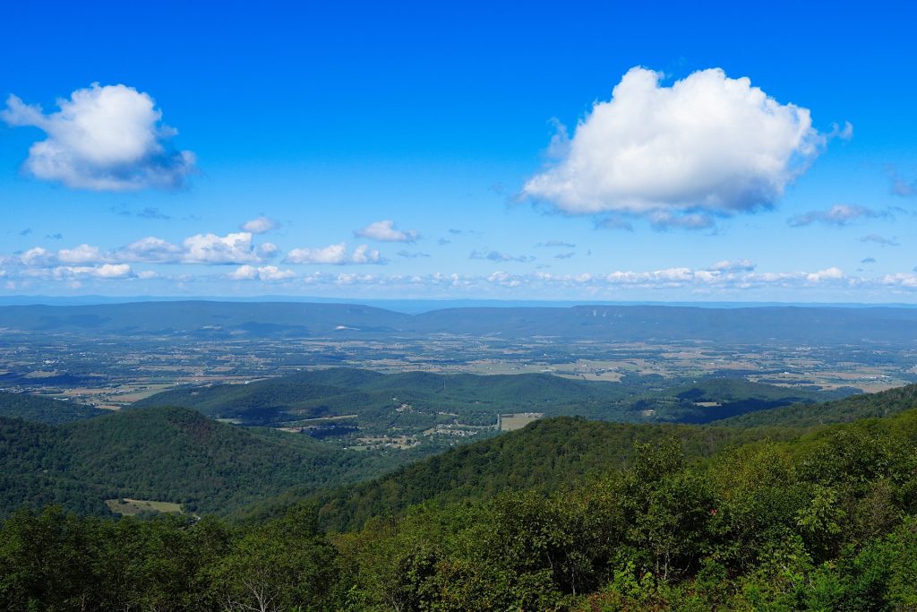 Scenic view of a valley surrounded by wooded hills under a clear blue sky with scattered clouds.