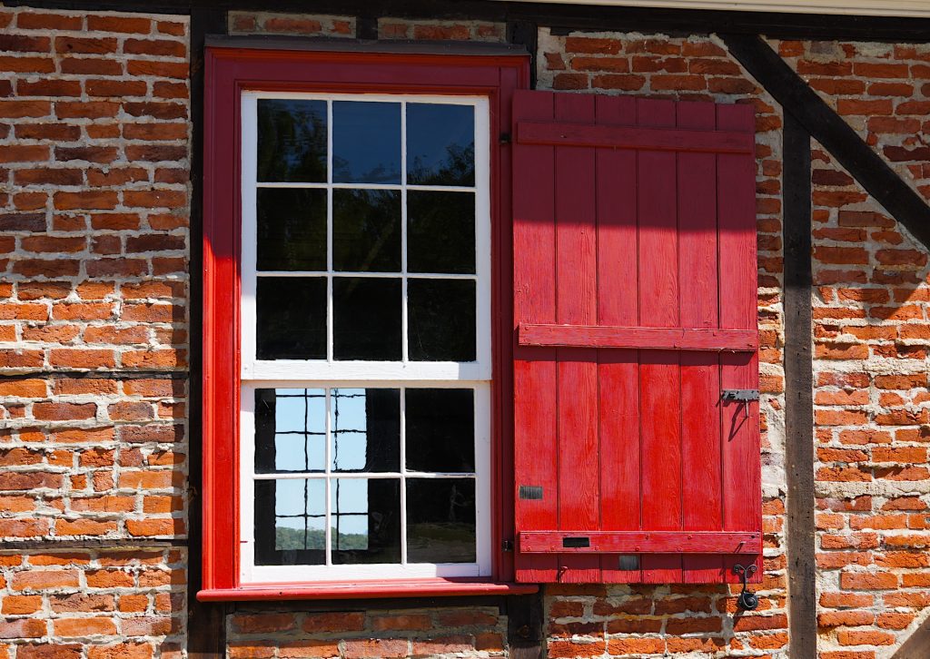 A red-framed window with a closed red shutter on a brick wall. The window has two sections with grid patterns, reflecting blue sky and dark tree silhouettes.