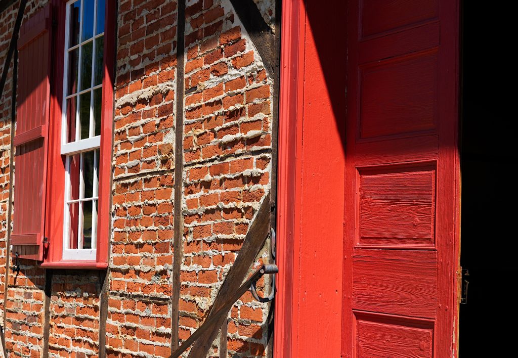 Red wooden door partially open on a brick building with a window and wooden beams.