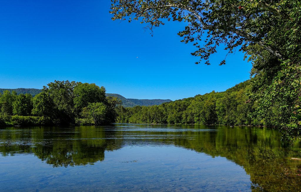 A calm river flows between lush green trees with distant hills under a clear blue sky.