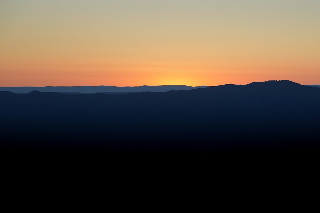 Sunset over a mountain range with a gradient sky transitioning from orange to blue, silhouetting the dark hills below.