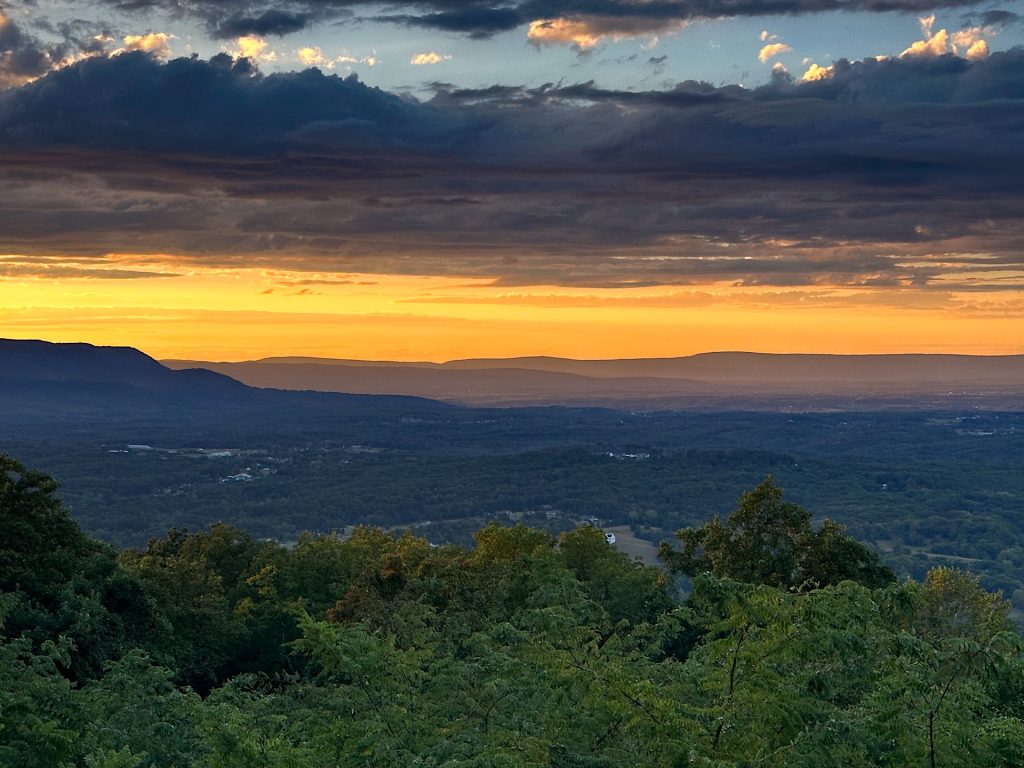 Golden sunset over a mountainous landscape with a foreground of lush green trees and a dramatic sky filled with scattered clouds.