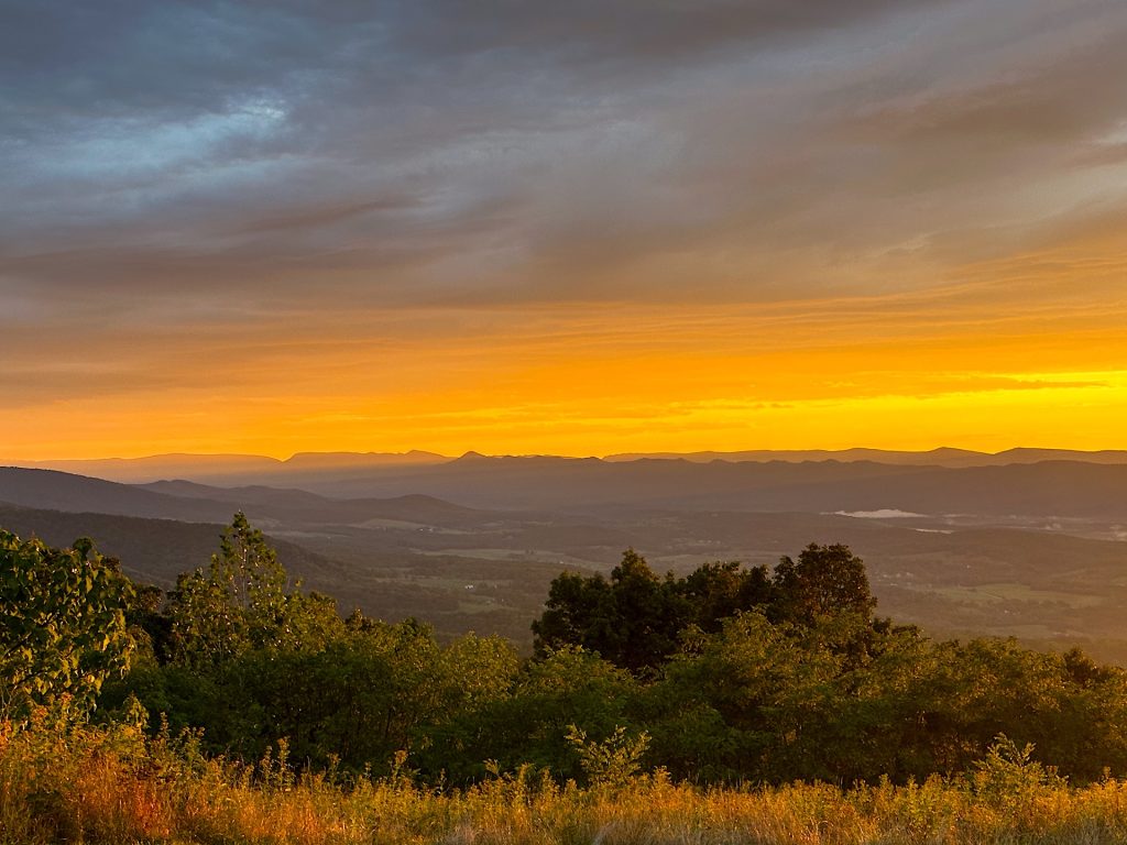 Sunset over a mountain landscape with vibrant orange and yellow hues in the sky, silhouetted mountain ridges in the distance, and lush green foliage in the foreground.