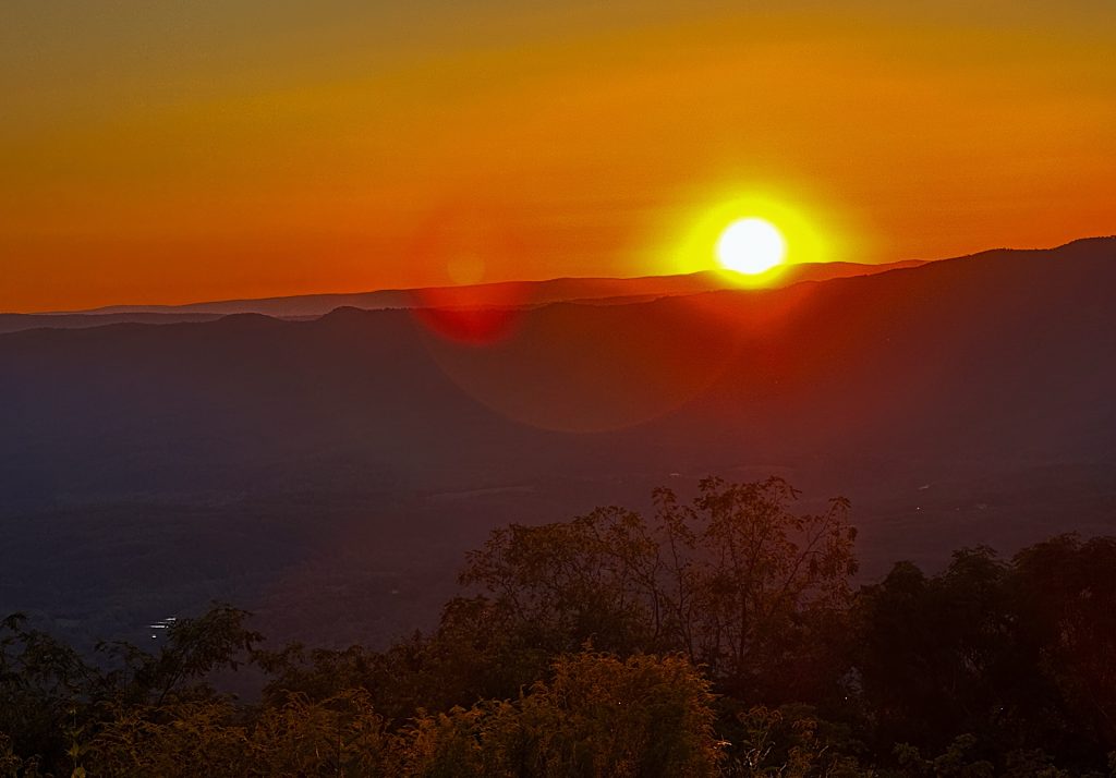 Sun setting over distant mountains, casting an orange glow in the sky, with silhouettes of trees in the foreground.