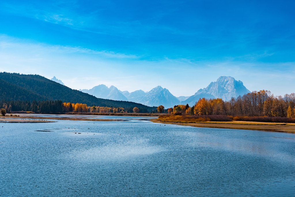 A serene lake with calm blue water, bordered by autumnal trees, with a distant mountain range under a clear blue sky.