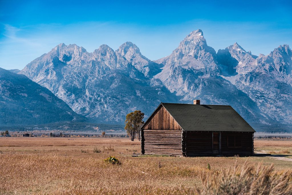 A wooden cabin stands in a vast field with towering mountains in the background under a clear blue sky.