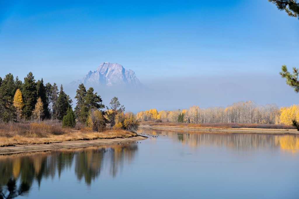 A tranquil river reflects trees with autumn foliage and a distant mountain under a clear blue sky.