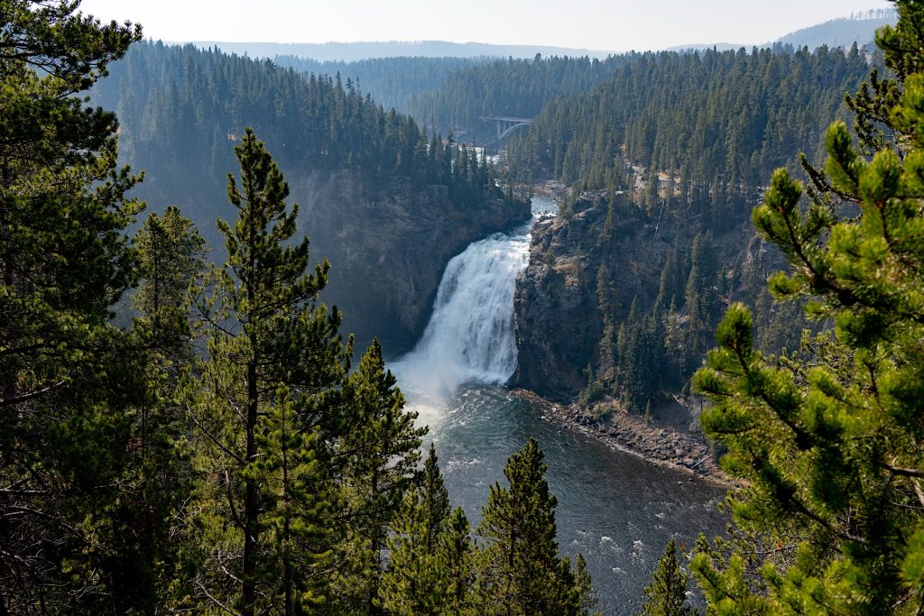 A waterfall cascades into a river surrounded by lush pine forests and distant hills, with a bridge visible in the background.