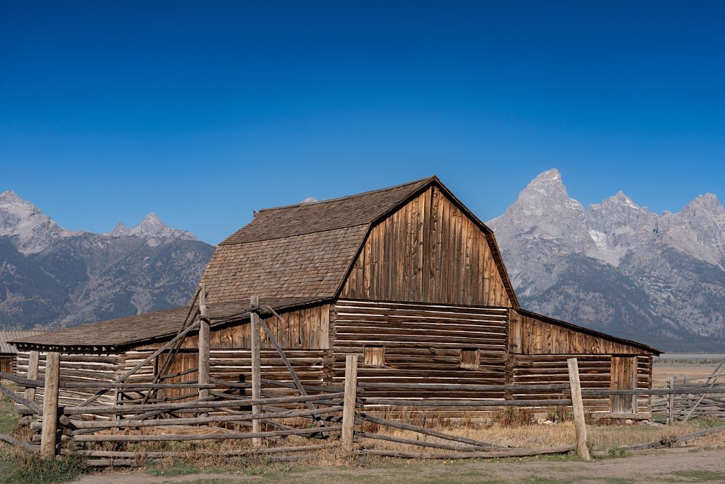 A rustic wooden barn with a shingled roof stands in front of a mountainous backdrop under a clear blue sky.