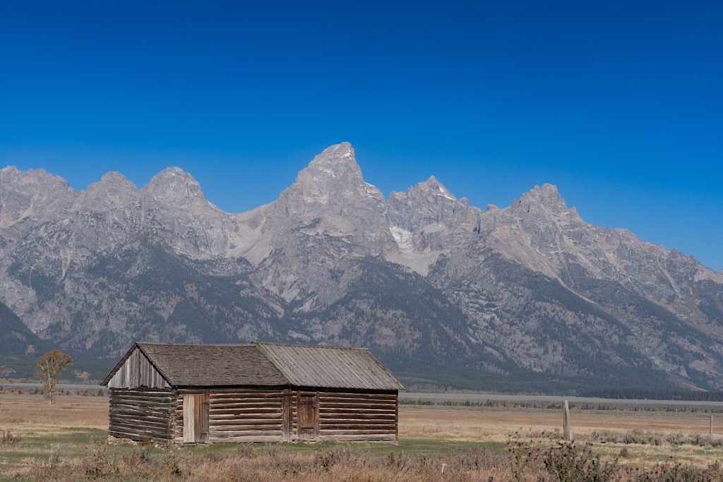 A rustic wooden cabin stands on an open field with towering mountain peaks in the background under a clear blue sky.