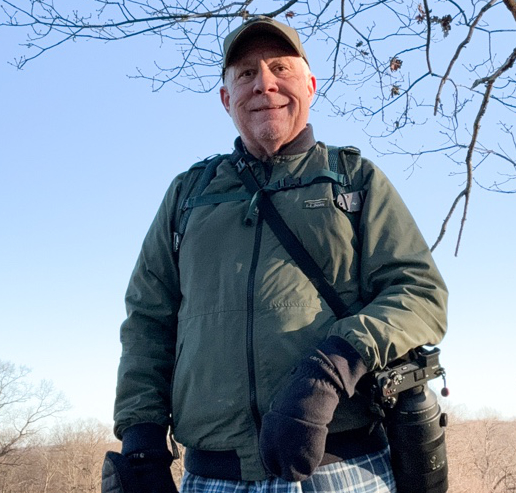 A person in a green jacket and cap stands outdoors, holding a camera with a large lens, supported effortlessly by their Peak Design harness. Bare tree branches and a clear sky form the background.