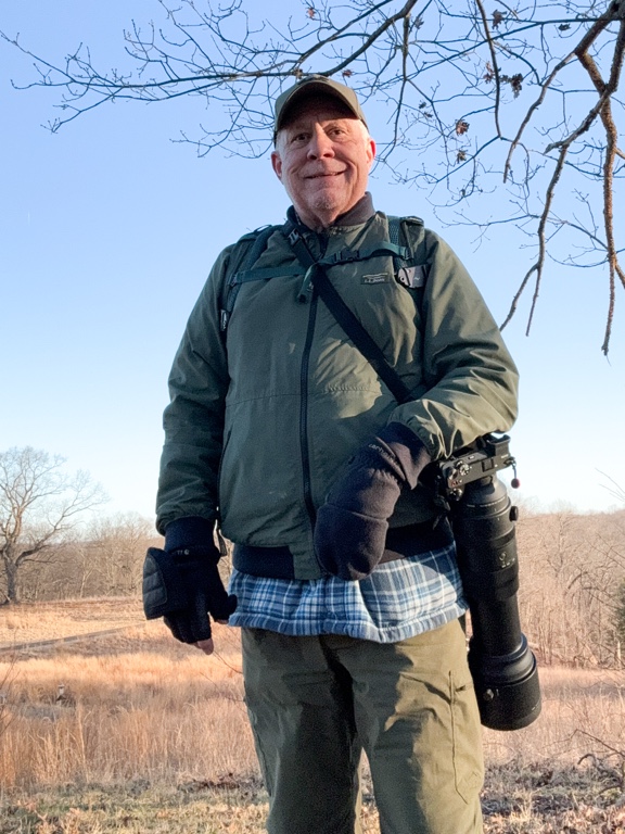 Person in outdoor gear stands in a field with a large camera lens, wearing gloves and a cap, tree branches overhead.