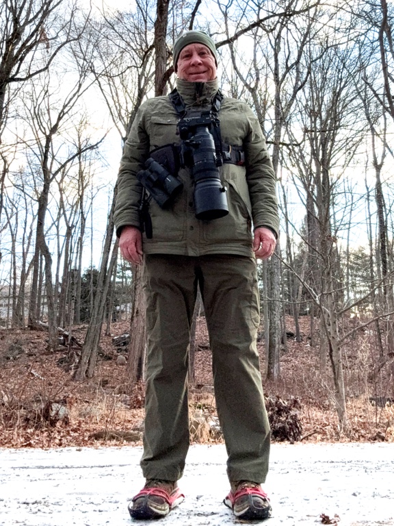 Person in outdoor clothing with a large camera lens, standing on a snowy path surrounded by bare trees.