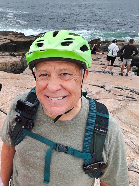 A smiling person wearing a green helmet and backpack, equipped with a Cotton Carrier for their camera, stands on rocky terrain near the ocean. Other people are in the background, taking in the breathtaking view.